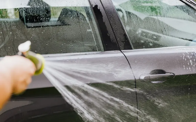 A person uses a hose to wash a black car outdoors, creating water splashes.