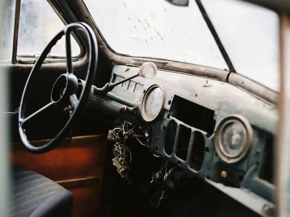 Interior of old fashioned automobile with seat and dirty windshield near big steering wheel and wire bundle above in daylight