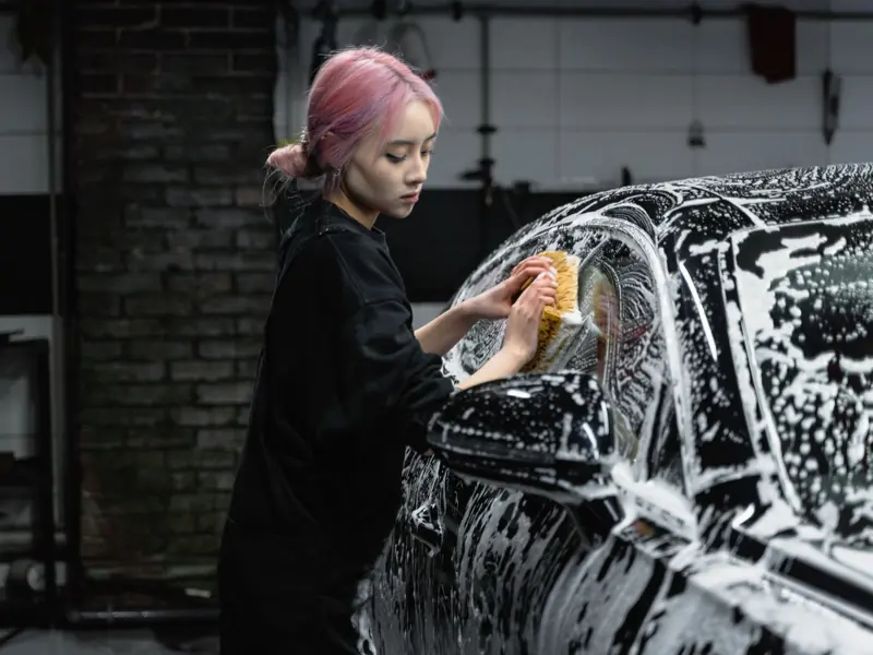 Woman with pink hair washing a car in a garage using soap and a sponge.