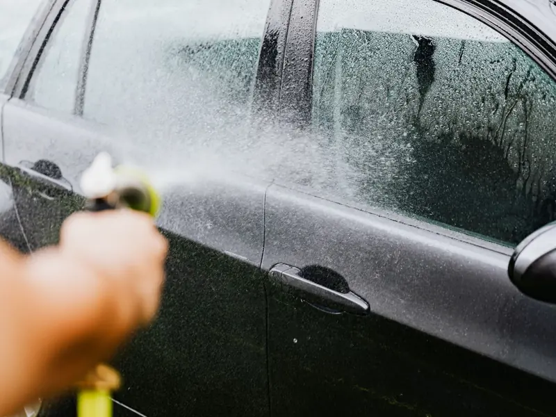 A car being washed with a hose outdoors. Water sprays create a cleaning effect on the vehicle.