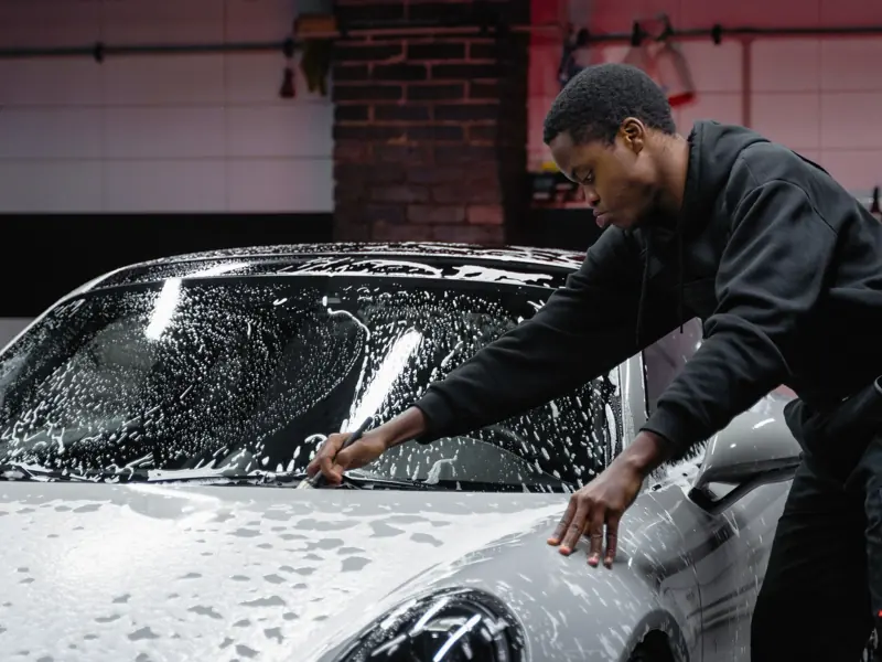 A man cleans a luxury sports car with soap in an indoor garage setting.