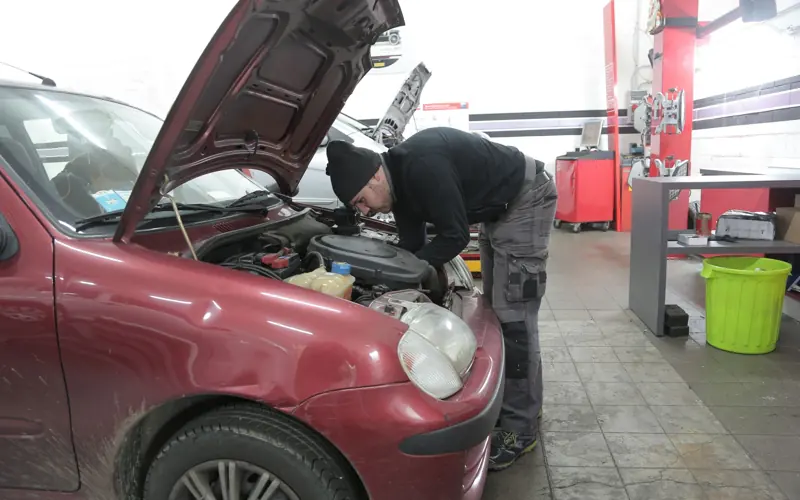 Mechanic performs engine inspection on a red car indoors, in a garage workshop.