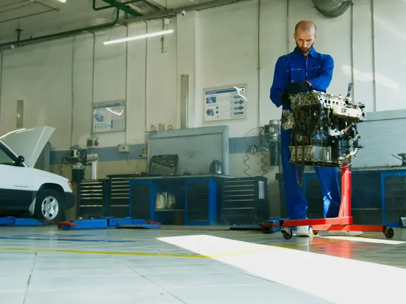 Mechanic lifting a car engine in a well-lit auto repair shop with vehicle in background.