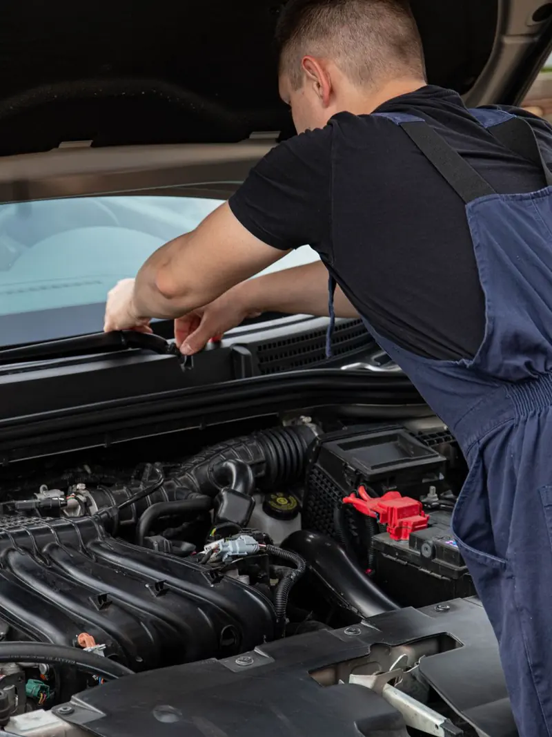 A mechanic in blue overalls works on a car engine outdoors, showcasing vehicle maintenance skills.