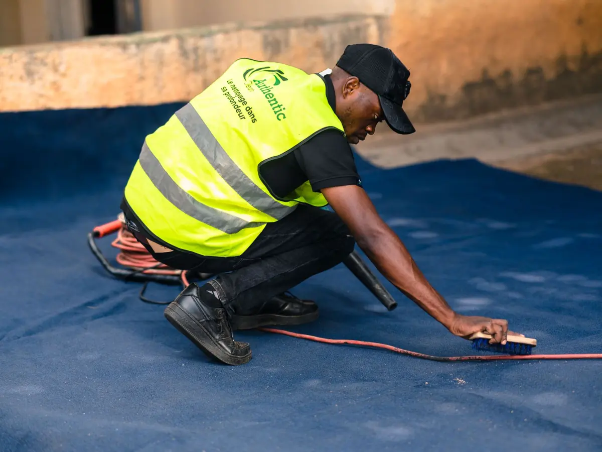 A worker dressed in a safety vest cleans a blue carpet outdoors with a brush.