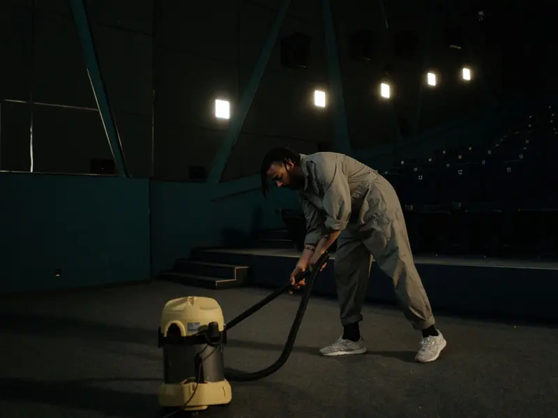 A cinema employee cleaning the theater floor with a vacuum cleaner under dim lights.