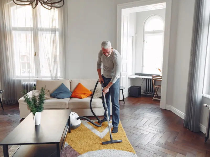 From above of focused grey haired man with beard vacuum cleaning carpet at home