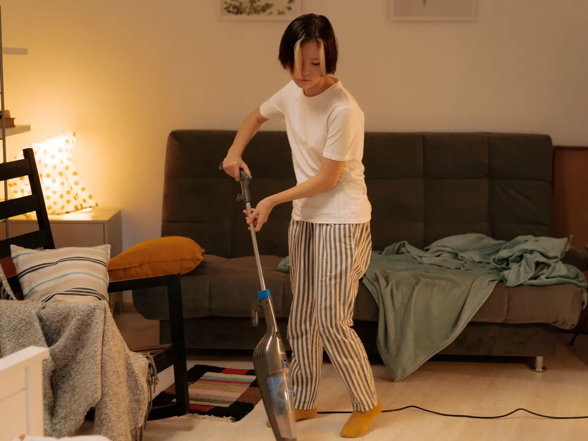 Asian woman cleaning a messy living room at home with a vacuum cleaner, creating a tidy space.