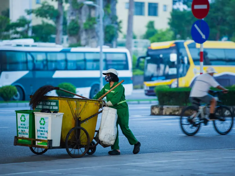 Street cleaner pushing cart with recycling bins in a busy urban setting.
