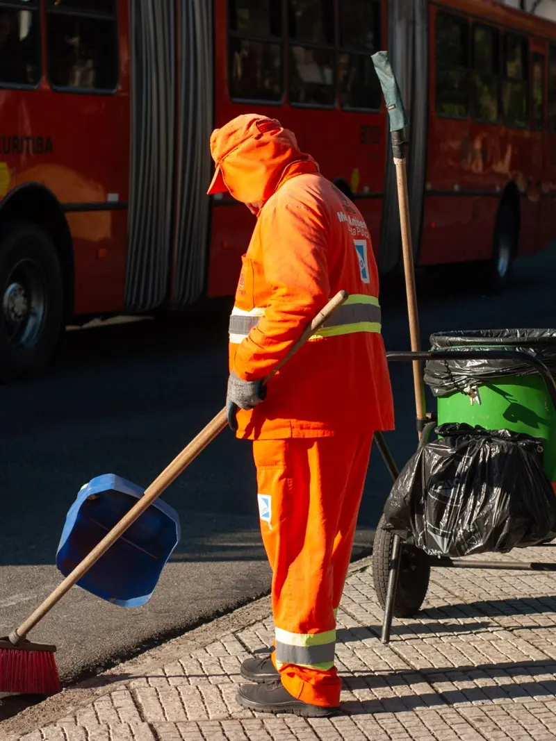 Street cleaner in bright orange uniform sweeping sidewalk near public bus in city setting.