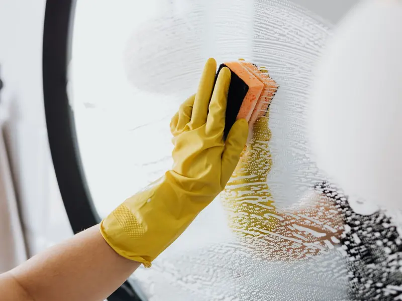 Anonymous person in yellow rubber gloves using simple sponge for cleaning mirror in bathroom