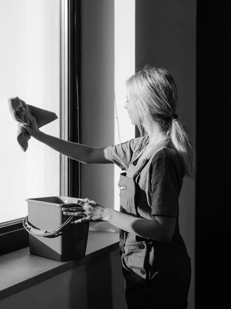 Black and white image of a woman cleaning a window with a cloth and bucket indoors.