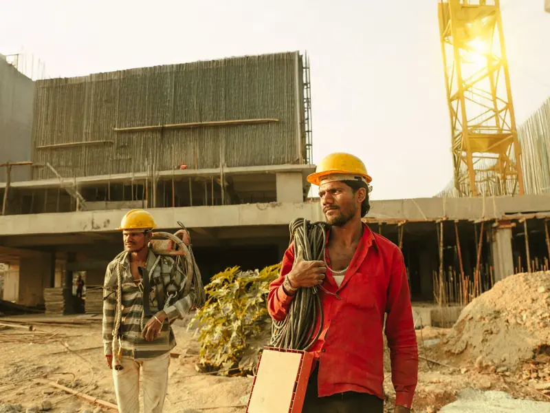Two construction workers wearing helmets at a site in India with a crane and building.