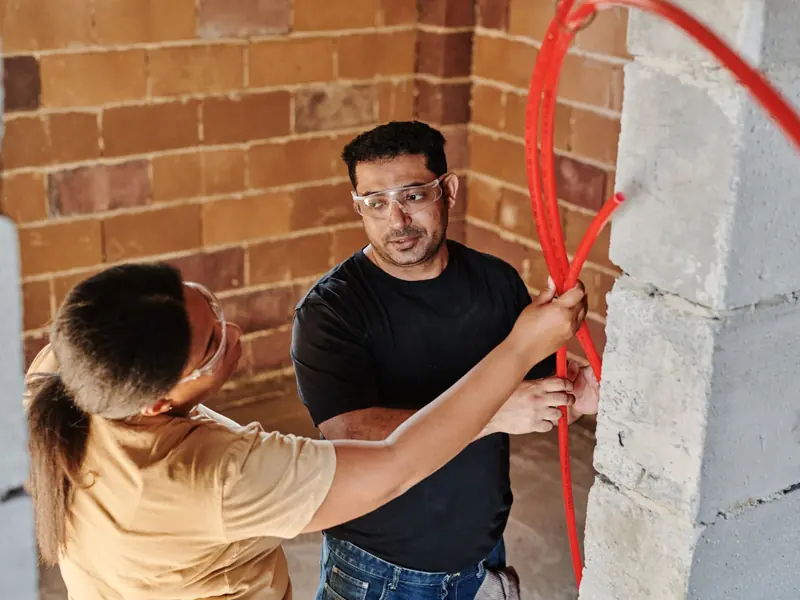 Two construction workers fitting red tubing on a brick wall at a construction site.