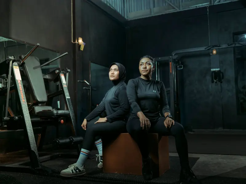 Two women in athletic wear sitting on a box with gym equipment in the background.