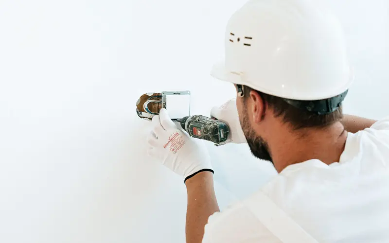 Construction worker wearing helmet and gloves uses a power tool to fix a wall.