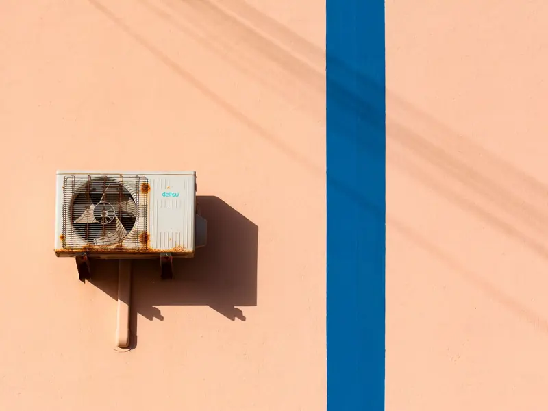 Rusty air conditioning unit casting shadow on a peach wall with blue stripe.