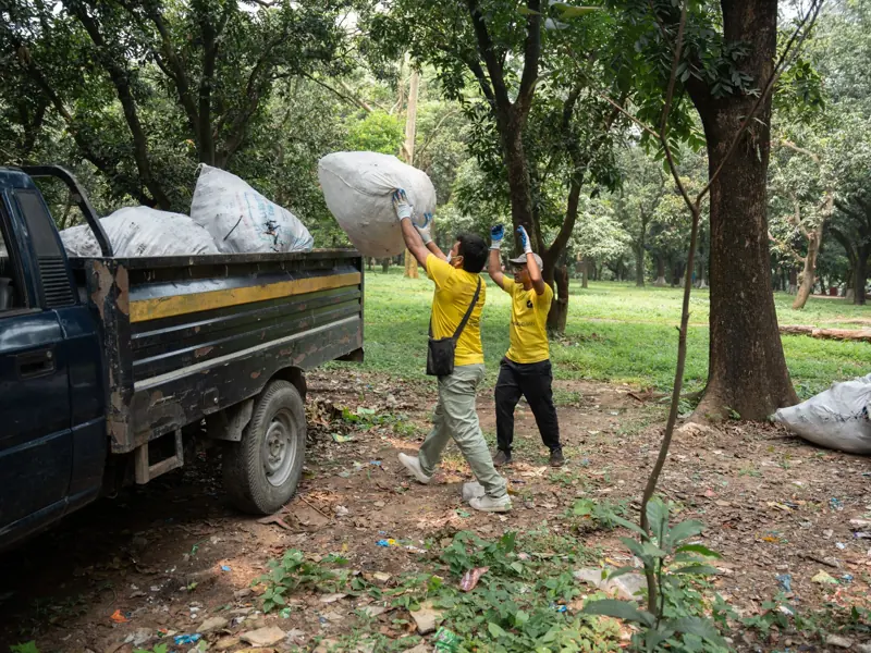Two workers in yellow shirts load a truck with large garbage bags in a wooded park area.