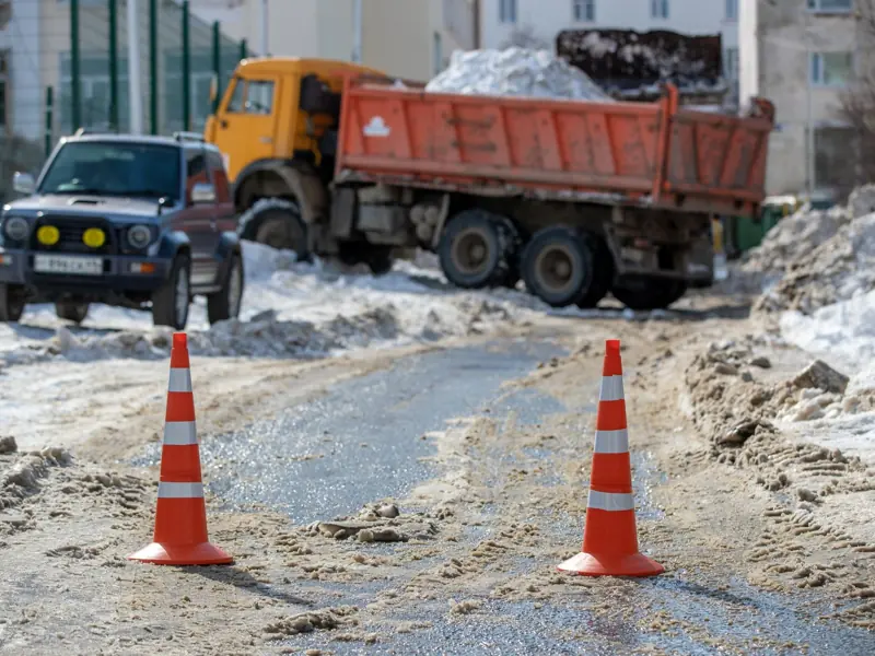 Orange traffic cones on a snowy road with parked vehicles and construction equipment in the background.