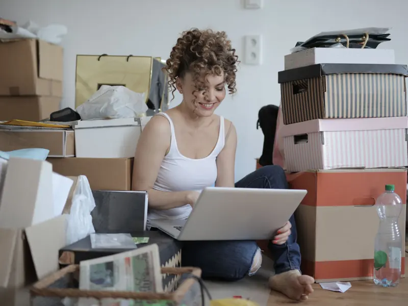 Smiling female customer in casual wear sitting on floor in untidy room with stacks of cardboard boxes and making orders in online store while using laptop