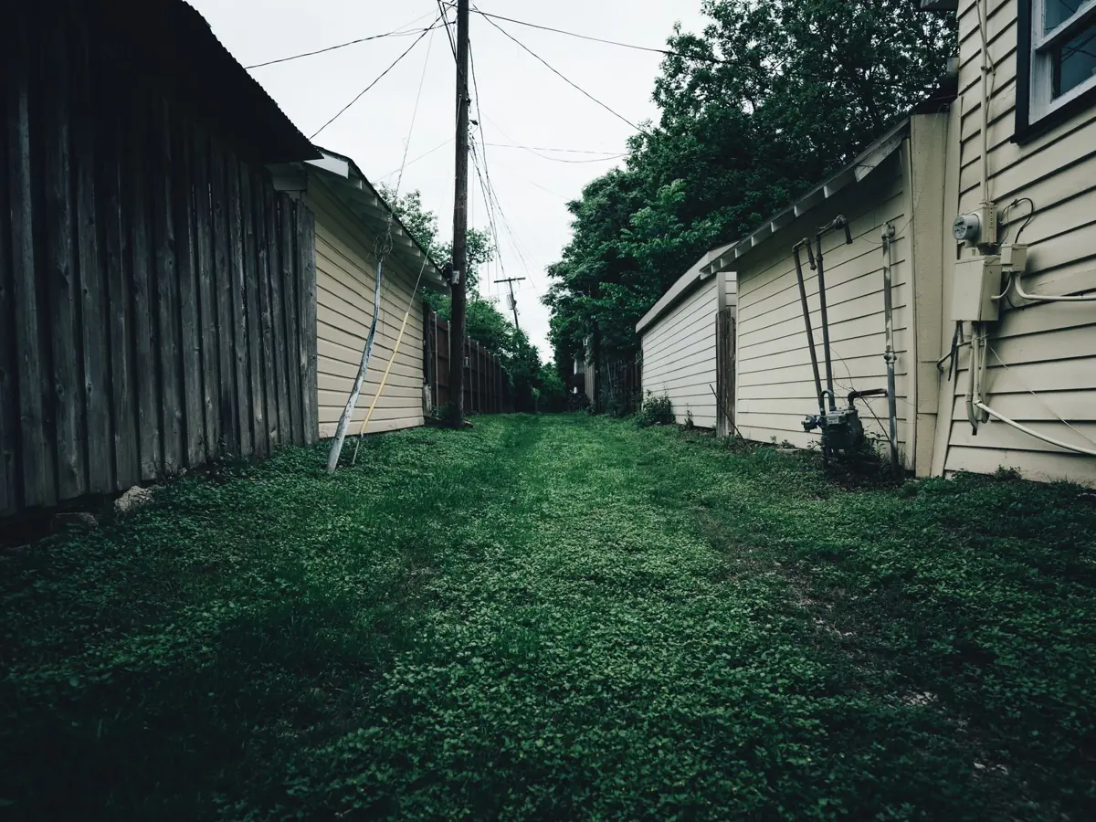 Rustic alleyway with grass pathway between wooden and metal fences in a residential area.