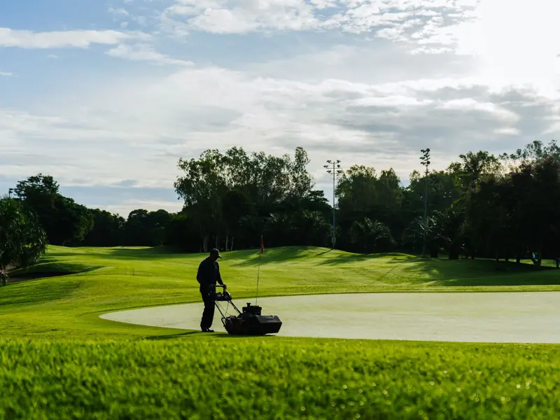 Groundskeeper mowing the green on a golf course during a sunny day, showcasing lush landscapes and maintenance.