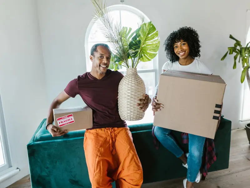 Couple smiling while moving into a new home, carrying boxes and a plant.