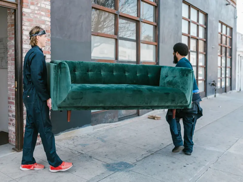 Two men carrying a green sofa on a city street, showcasing teamwork and moving services.