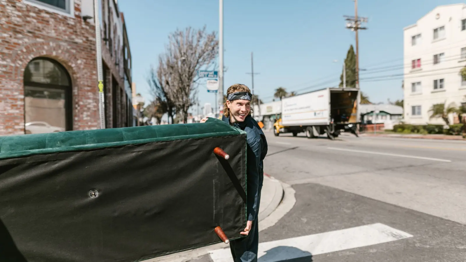 A cheerful man carries a large couch across a sunny urban street during a move.