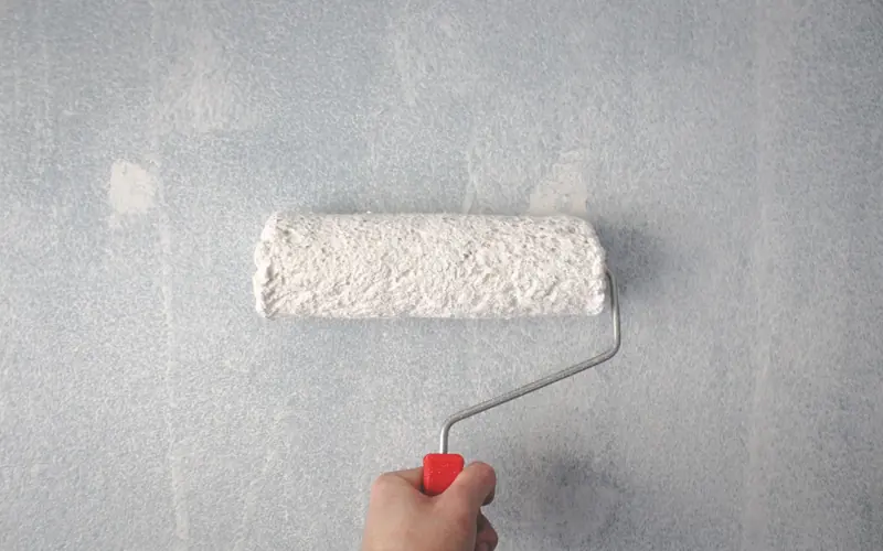 A person's hand using a paint roller to apply white paint on a wall.