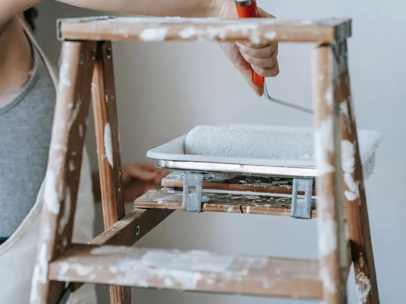 Adult woman using a roller and ladder to paint a room wall indoors.