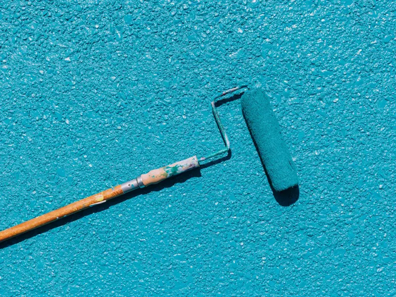 A close-up of a blue paint roller applying paint on a textured wall surface, captured in bright sunlight.