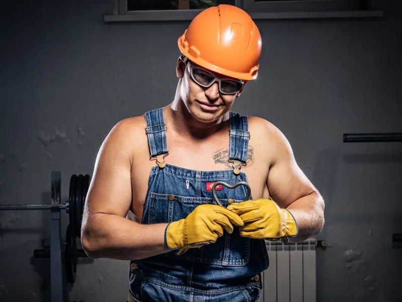 Muscular construction worker in overalls and helmet displaying strength and humor indoors.