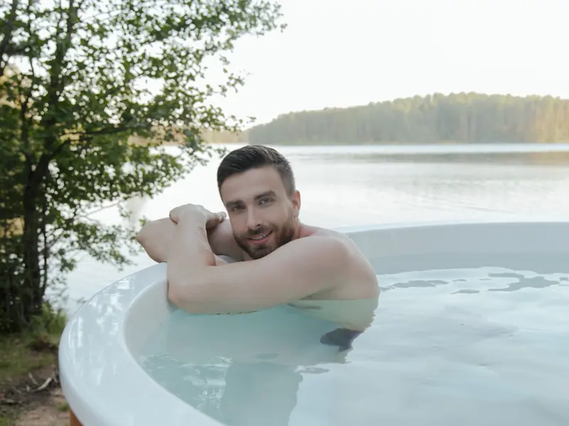 Shirtless man enjoying relaxation in hot tub by scenic lake, smiling and embracing leisure outdoors.