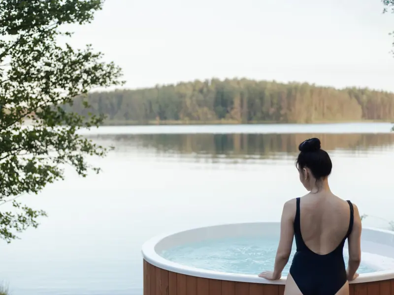 A woman in a swimsuit relaxes in an outdoor hot tub by a serene lake surrounded by trees.