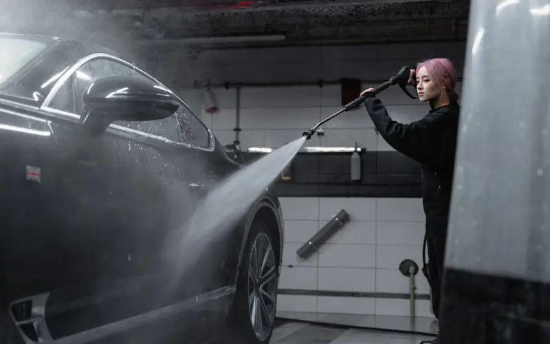 Woman power washing a black car in an indoor garage using a high-pressure washer.