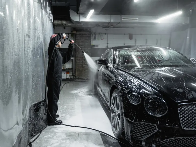 A person in a garage power washing a black car covered in soap and water spray.