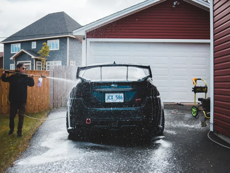 Full body of young male in black wear using long thin hosepipe while washing luxury vehicle in yard of tall private house
