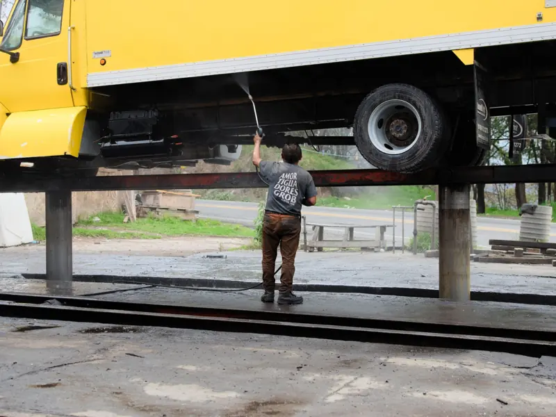 A worker power washes the underside of a raised yellow truck at an outdoor service station.