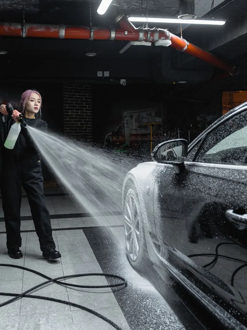 Woman using a pressure washer to clean a black car in an indoor car wash setting.