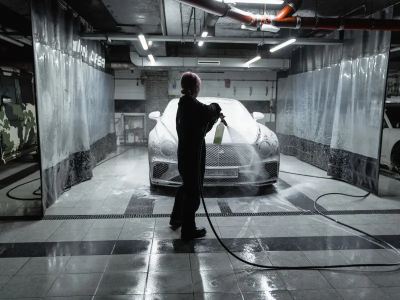 A person using a hose to spray a car with snow foam in an indoor carwash setting.