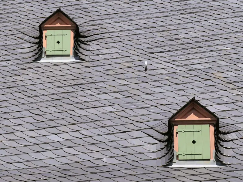 Close-up of a slate rooftop featuring two green shuttered dormers under daylight.