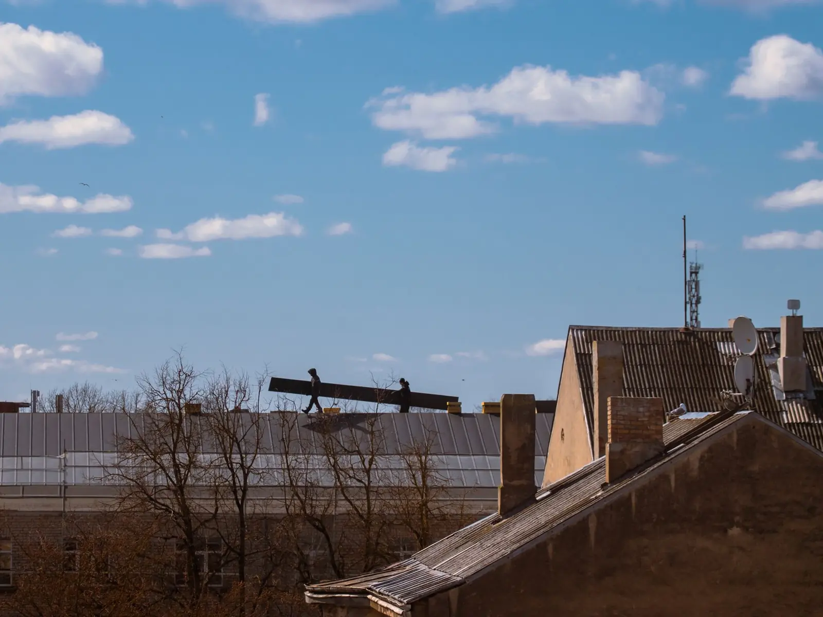 Silhouette of construction workers on a roof under a bright blue sky with clouds.