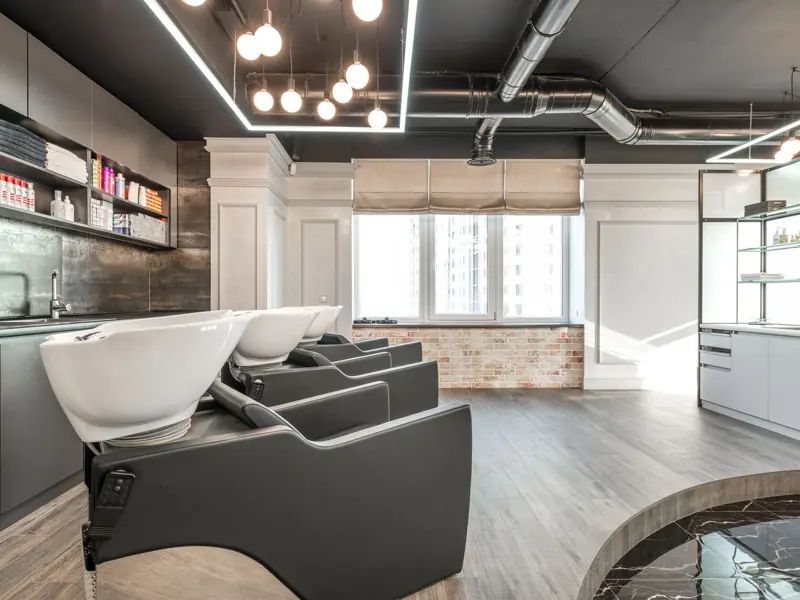 Black backwash chairs with sinks placed in modern spacious beauty salon with window and arrangement of cosmetology supply on shelves