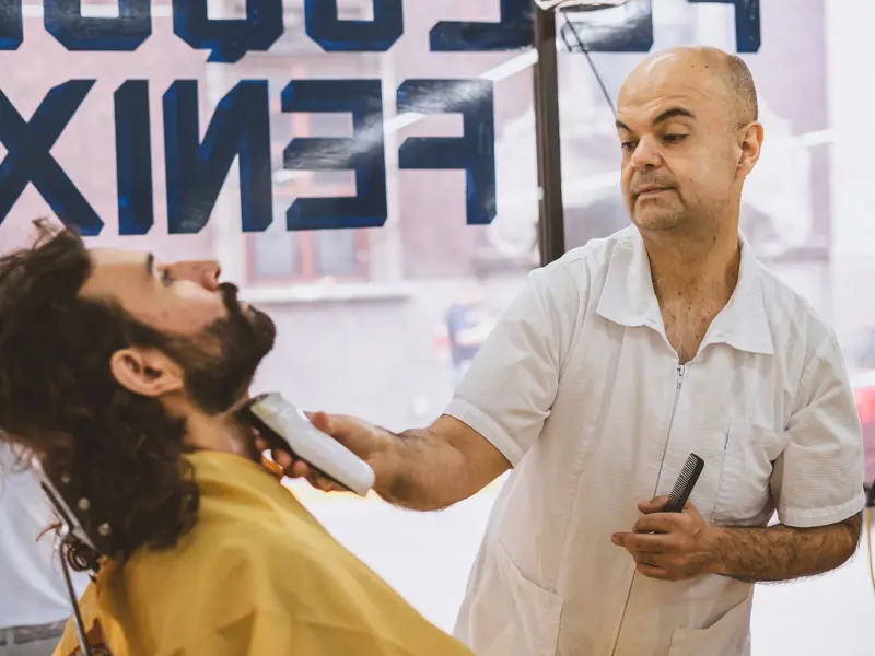 Barber expertly shaving a bearded client in a bustling barbershop.