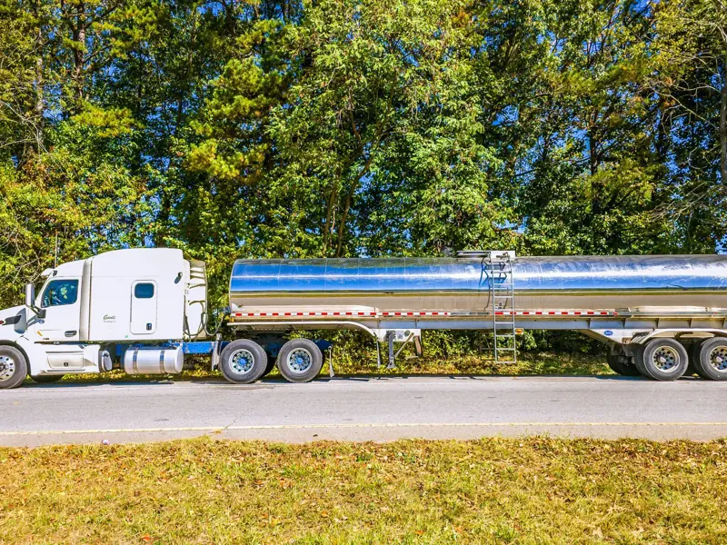 A modern white semi-truck with a polished tanker trailer parked on a roadside in a forested area during the day.