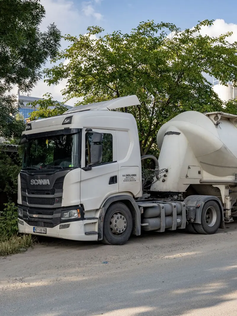 White Scania tanker truck parked by trees on a sunny city street.