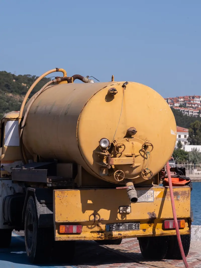 A yellow tanker truck parked by the harbor in Foça, İzmir, with a scenic coastal view.