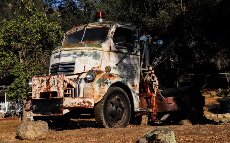 A rusty, abandoned tow truck sits outdoors, surrounded by nature and vintage charm.