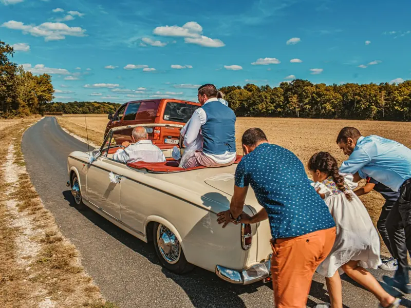 Family pushes classic wedding car in countryside, a humorous moment on sunny day.